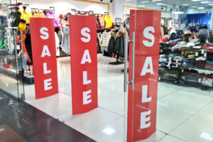 Entrada de loja de roupas com painéis vermelhos exibindo a palavra “SALE” em letras brancas, indicando promoção.