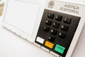Close na urna eletrônica brasileira com teclado numérico e botões branco, corrige e confirma, sob o título Justiça Eleitoral.