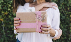 Mulher segurando um presente embrulhado em papel kraft com faixa rosa e laço de barbante, enquanto recebe um abraço por trás ao ar livre.