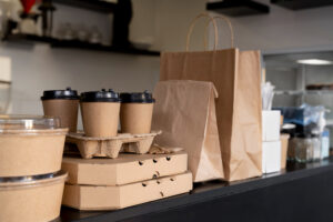 Embalagens de delivery com cafés, sobremesas e sacolas kraft organizadas sobre balcão de cafeteria.