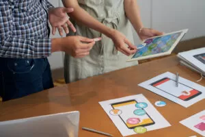 Duas pessoas analisam ícones de aplicativo em uma mesa com papéis e tablet, em um ambiente de trabalho colaborativo.