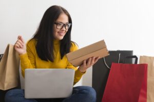 Mulher sorridente segurando uma caixa de encomenda enquanto usa um notebook, rodeada por sacolas de compras, representando compras online.